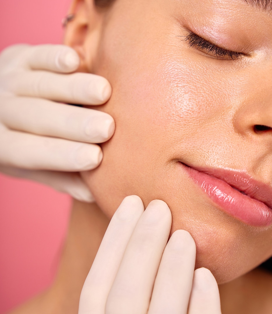 A close-up of a woman’s face with light makeup, showing a practitioner’s gloved hands gently examining her cheek. The background is pink, adding a soothing atmosphere.