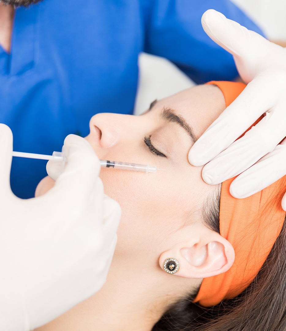 A medical professional in gloves performing an injection on a woman's face, who is lying back with her eyes closed. The woman is wearing a headband and earrings.