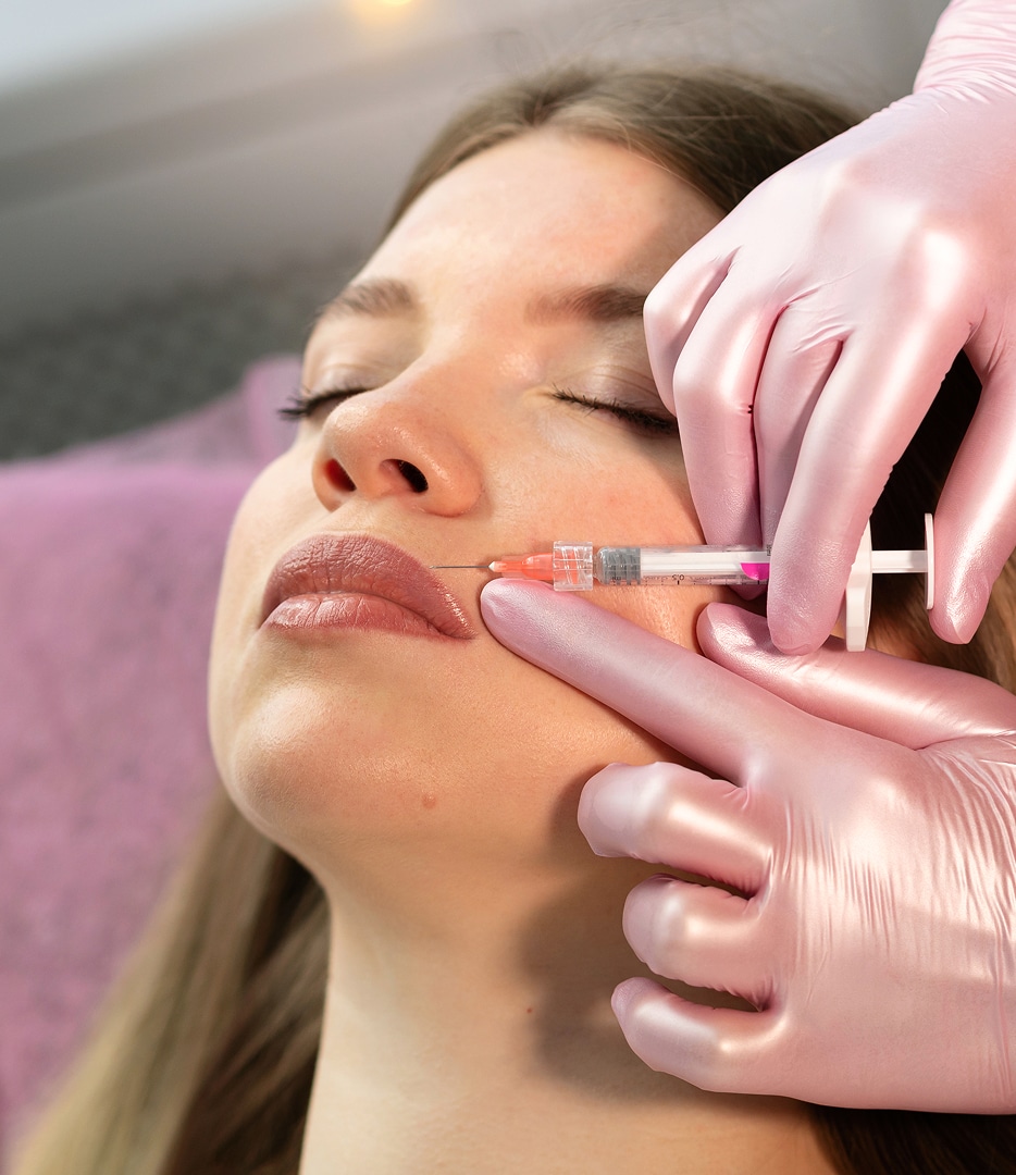 A close-up of a woman receiving a cosmetic injection. A healthcare professional in pink gloves holds a syringe near the woman's lips, who is relaxed with eyes closed. The background shows a medical or spa setting.