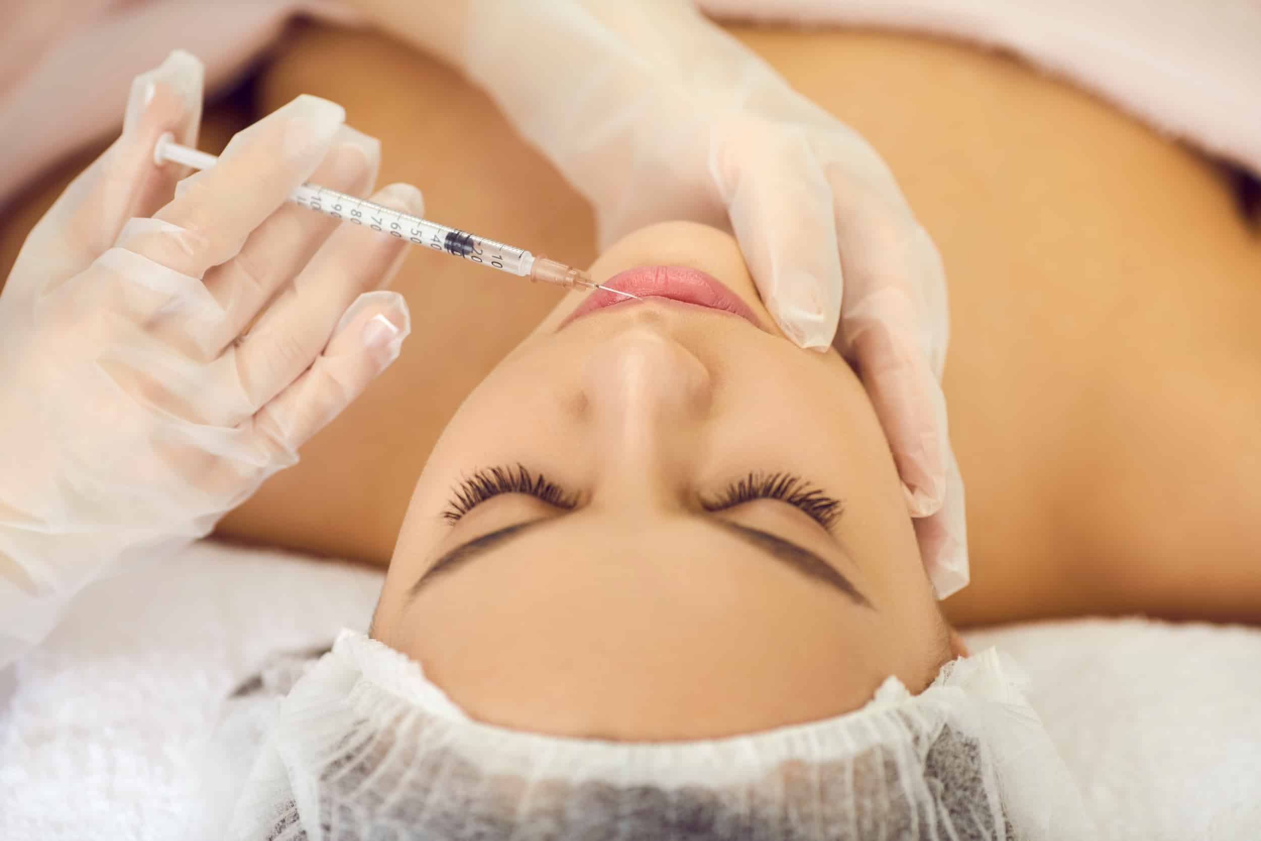 A woman receiving a cosmetic injection, lying on a treatment table. A professional wearing gloves is administering the injection to her lips, while the setting appears clinical and clean.