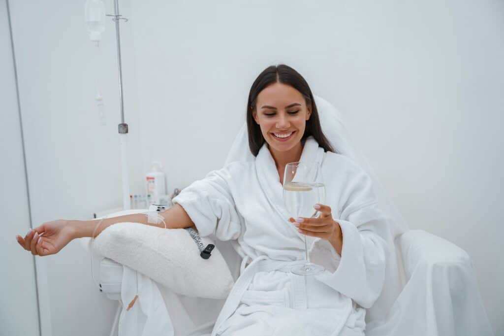 A woman wearing a white robe sitting in a chair, holding a glass of drink while receiving an IV treatment. The background is minimalistic with neutral colors and a calm atmosphere.