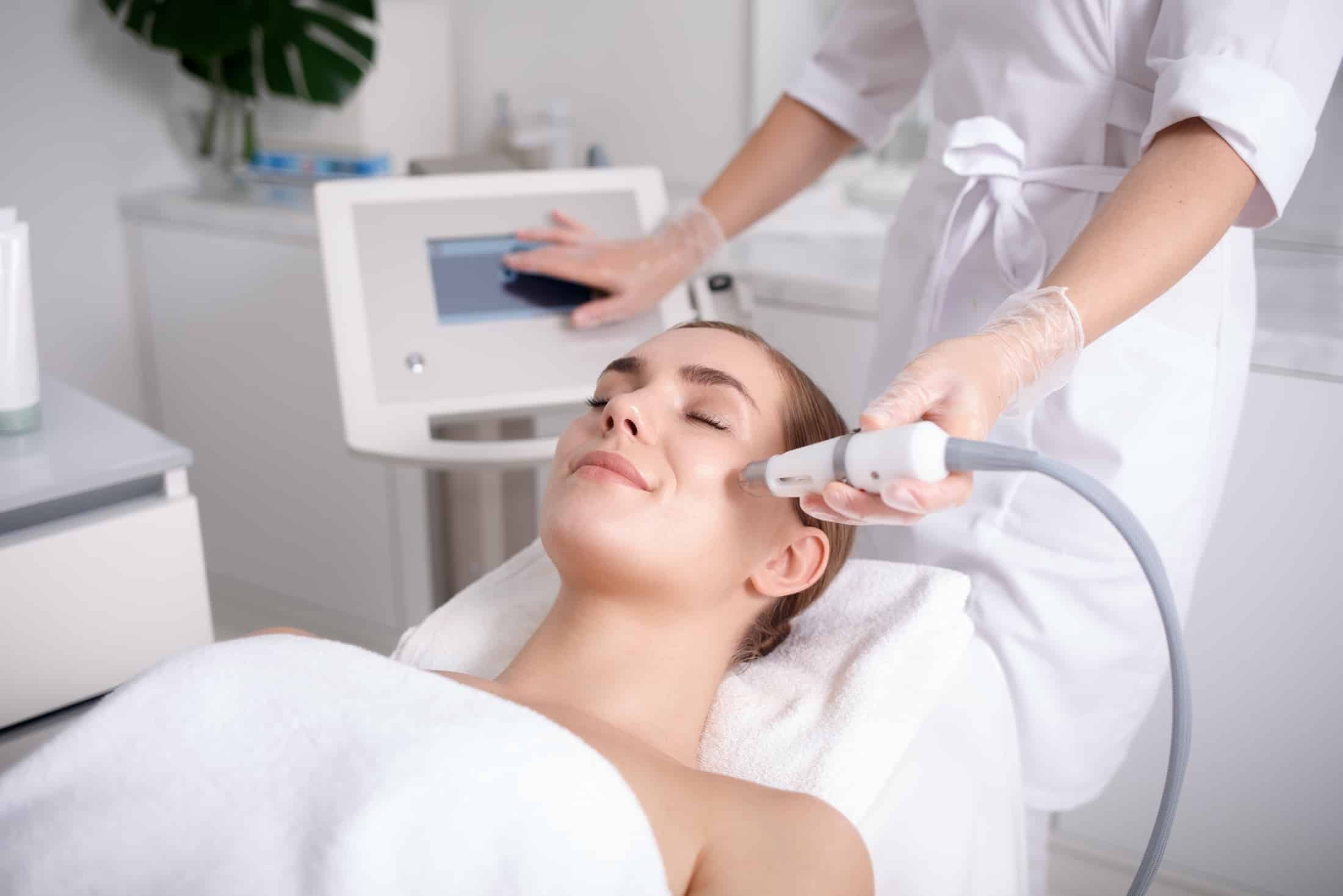 A woman receiving a facial treatment in a spa setting. A technician is using a device on the woman's face, while she relaxes on a treatment table. The environment is bright and modern with a plant in the background.