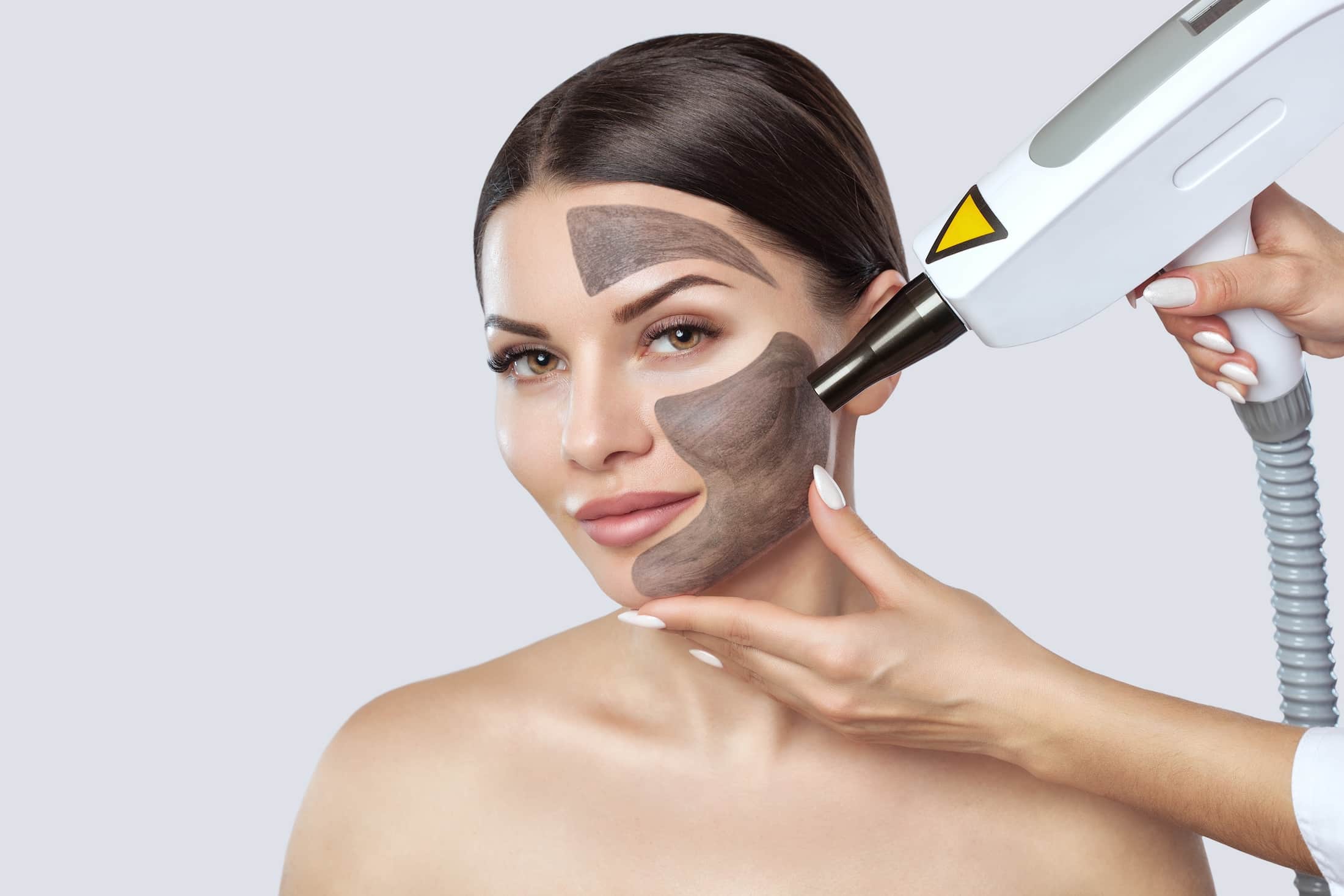 A woman receiving a facial treatment with a device, featuring dark mask-like sections on her face. A professional's hand is using the treatment applicator near her cheek. The background is a light gray color.