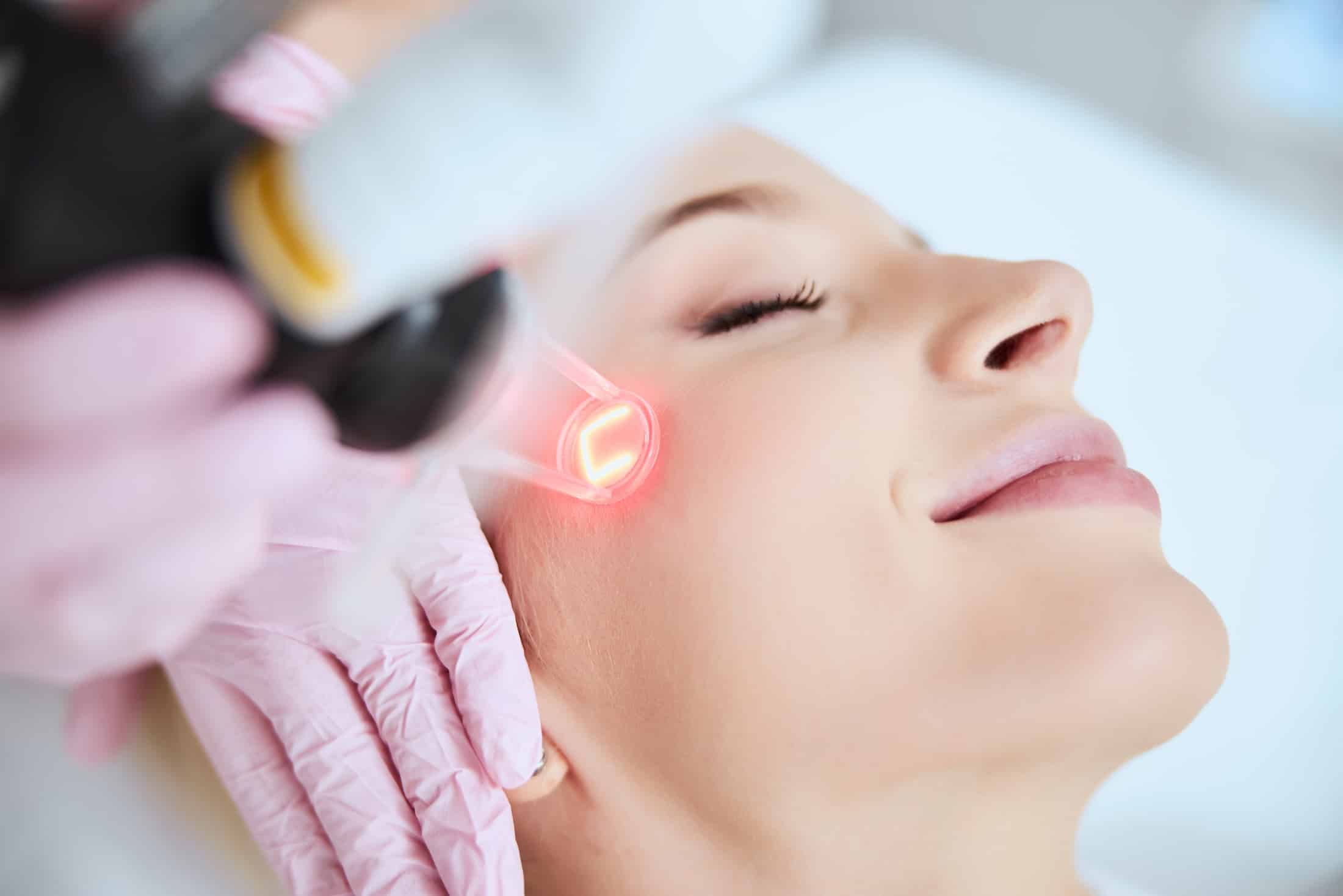 A woman receiving a facial treatment with a laser device, focused on her cheek. A professional wearing pink gloves operates the device, while the woman appears relaxed with her eyes closed.