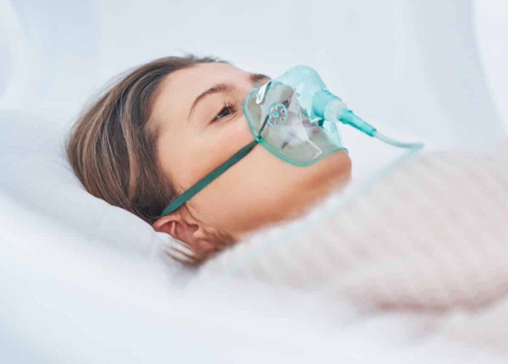 A woman lying on a hospital bed, wearing an oxygen mask. The focus is on her face, with soft lighting highlighting her features and the mask.