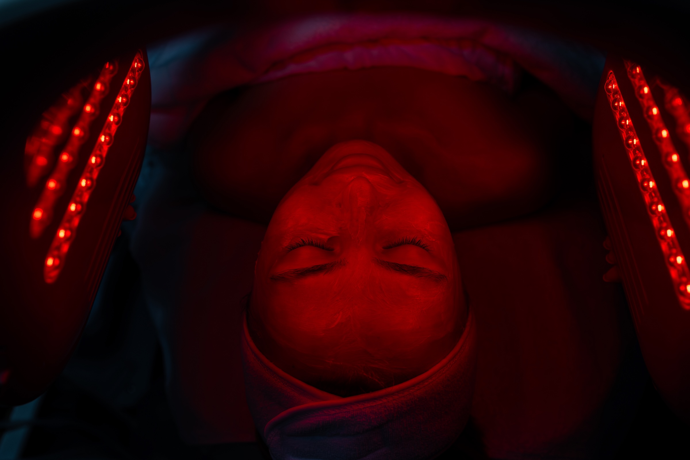 A person lying down under red light therapy devices, with a calm expression. The setting is dimly lit, highlighting the red lights on both sides of the face.