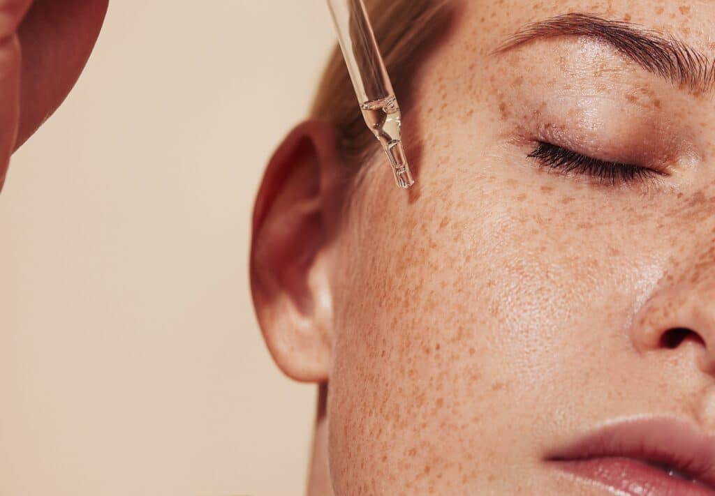 A close-up of a woman receiving a skincare treatment, with a pipette applying serum to her cheek. Her eyes are closed, showcasing a natural look with freckled skin and a soft expression.