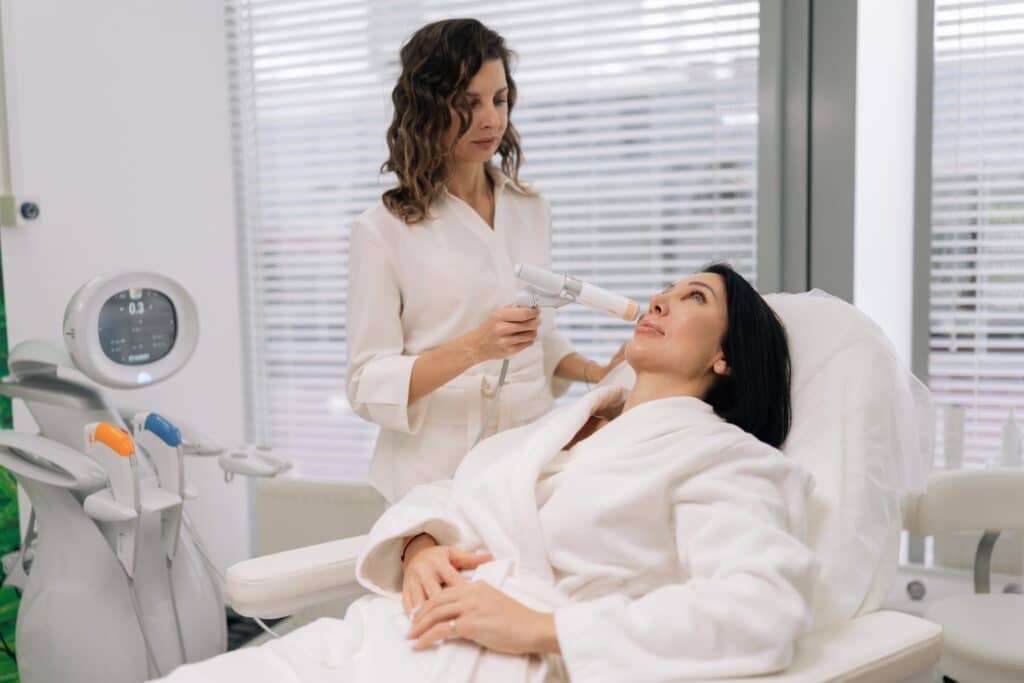 A woman in a white robe receives a skincare treatment while lying on a treatment bed. Another woman, wearing a white outfit, uses a handheld device on her face in a modern clinic setting.