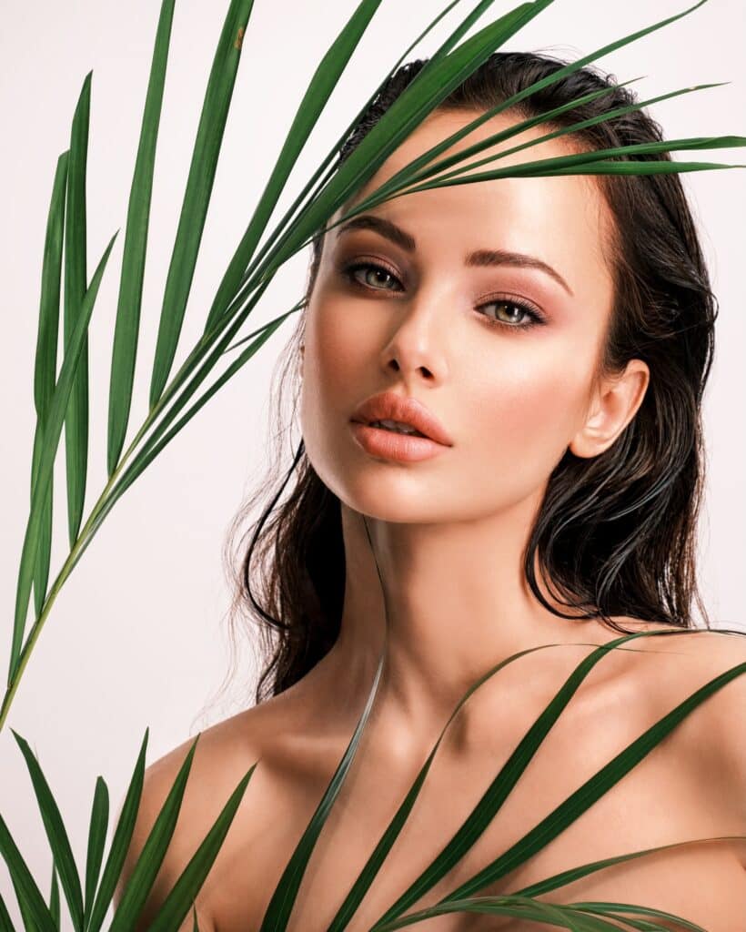 A woman with long, dark hair poses against a light background, partially concealed by green palm leaves. She has a neutral expression, focusing on the camera.