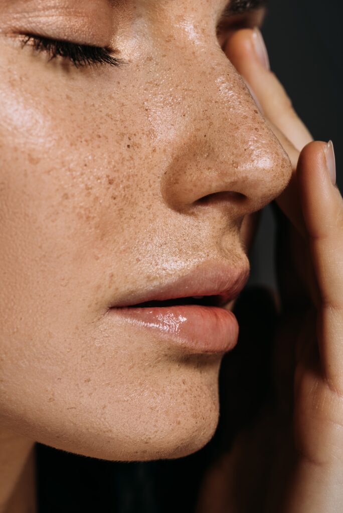 A close-up of a woman's face, highlighting her freckles and natural texture. She is touching her cheek gently with one hand, and her eyes are closed, indicating a calm expression.