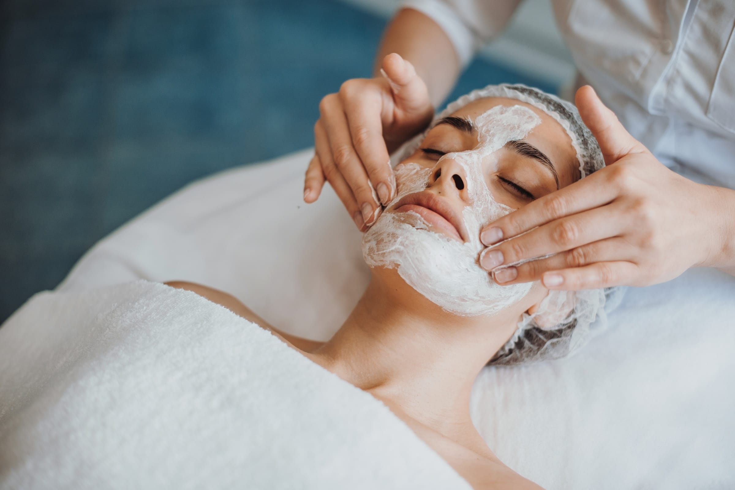 A woman receiving a facial treatment, with a therapist applying a white cream mask on her face. The client is lying on a spa bed, covered with a white towel, while the setting appears tranquil with soft lighting.