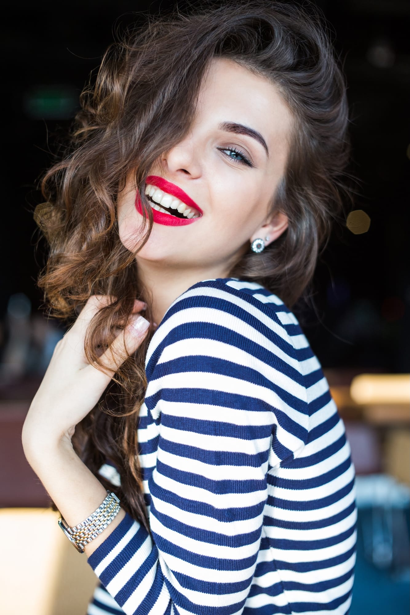 A woman with long, wavy hair wearing a striped navy and white shirt, smiling brightly with bold red lipstick. She appears to be in a casual setting with soft lighting in the background.
