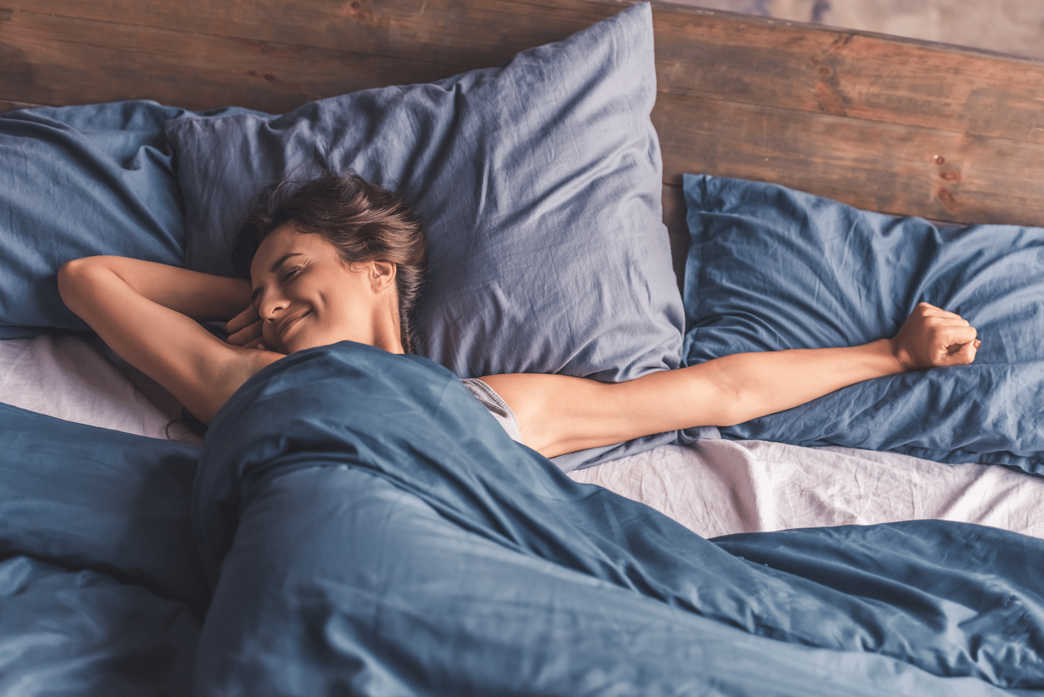 A woman stretching comfortably in bed, surrounded by blue bedding and pillows. She has a relaxed expression, with her arm raised above her head and a slight smile on her face, suggesting a peaceful morning or restful moment.