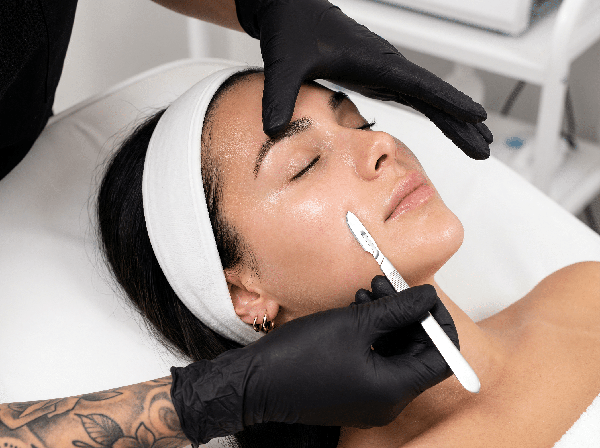 A person receiving a skincare treatment, lying on a treatment bed. A specialist, wearing black gloves, uses a facial tool on the person's skin. The person has a headband on and their eyes are closed, with a clean, minimalist treatment room in the background.