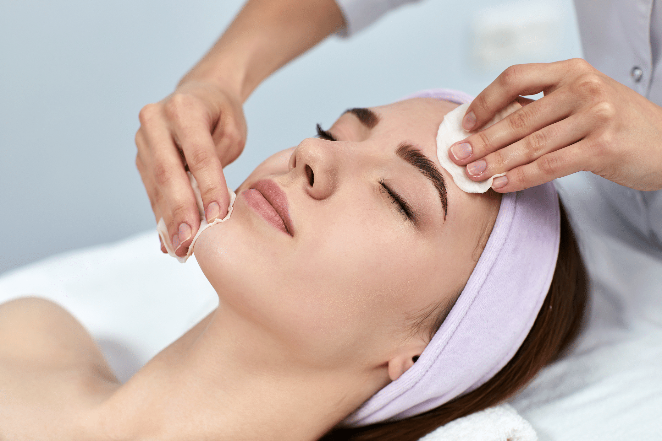 A woman receiving a facial treatment. She is lying on a massage table with her eyes closed, wearing a light purple headband. A technician is using a cotton pad on her cheek, applying a skincare product in a tranquil spa setting.