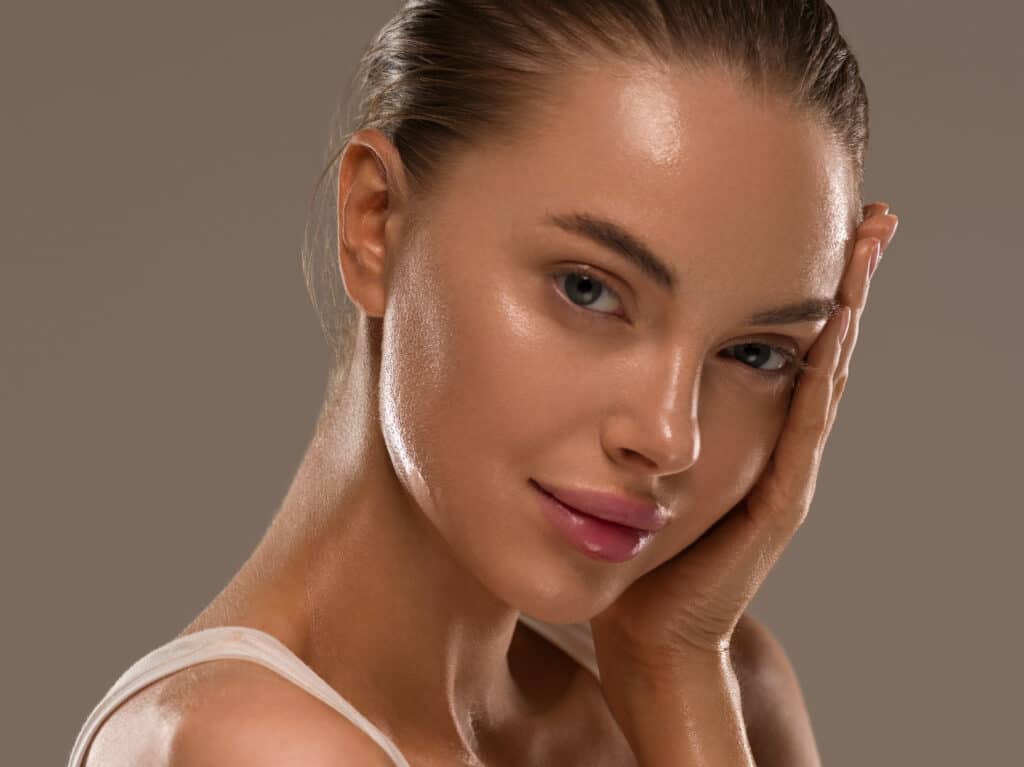 A close-up portrait of a woman with fair skin, softly illuminated. She is wearing minimal makeup, showcasing a natural look. One hand rests on her cheek, and she is gazing slightly towards the camera with a relaxed expression.