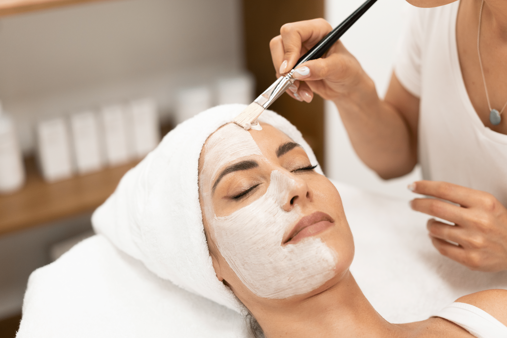 A woman lying on a treatment table in a spa, receiving a facial treatment. A professional is applying a white facial mask with a brush. The woman has a towel wrapped around her head and appears relaxed, with her eyes closed.