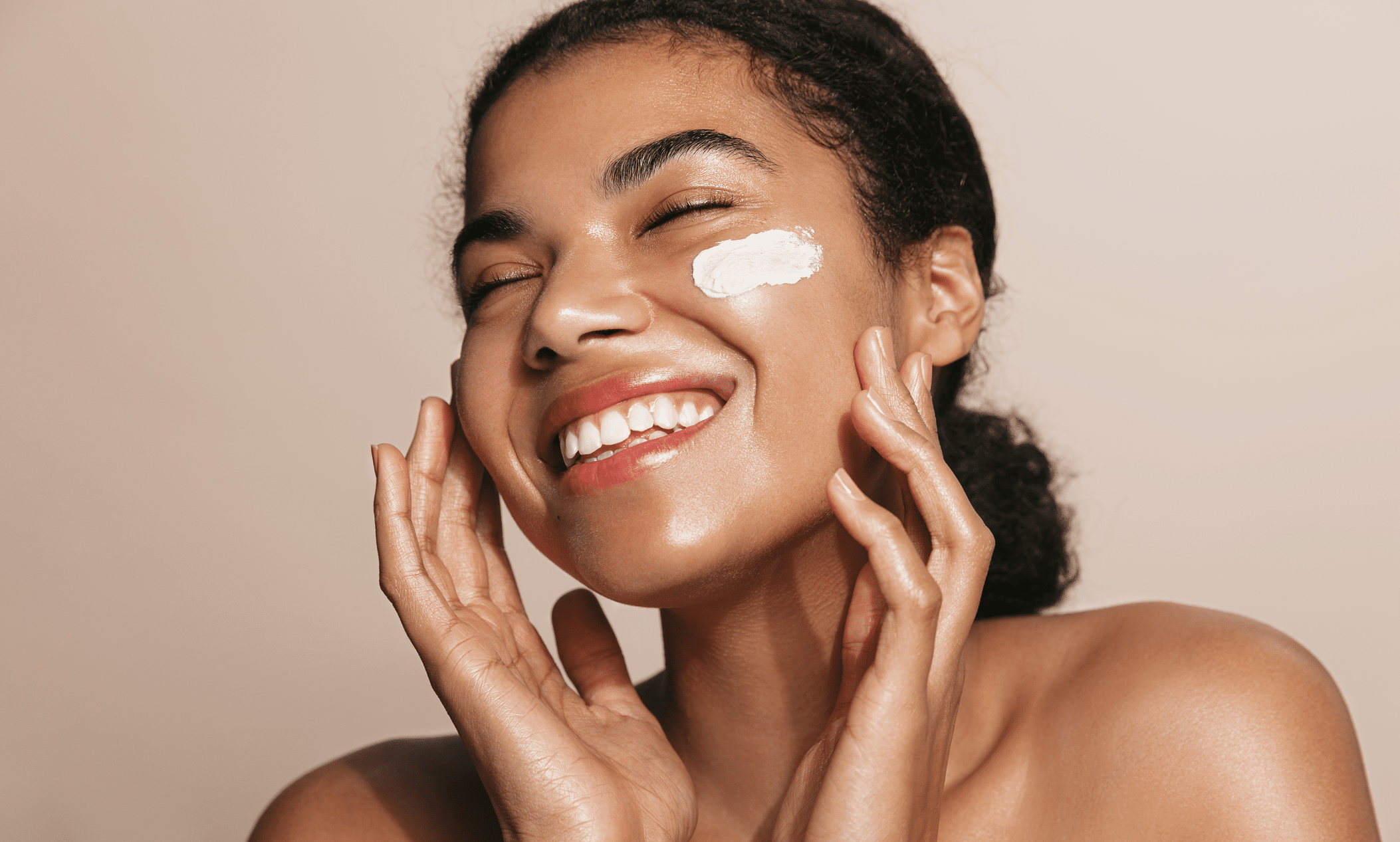 A smiling woman with dark, curly hair applies cream to her cheek. She has a light application of moisturizer on her face and is posing against a soft beige background. Her expression conveys enjoyment and confidence.