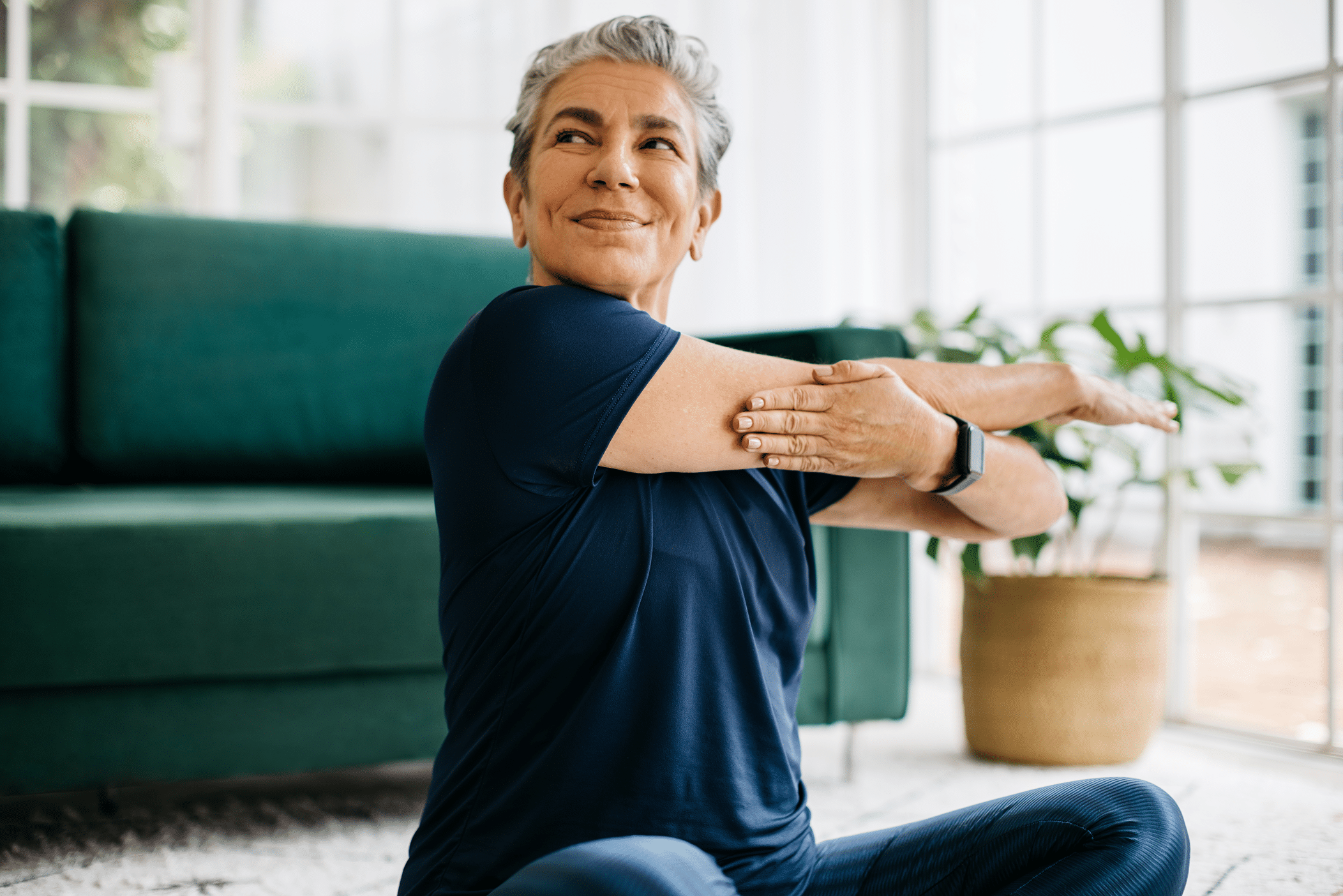 A woman with gray hair in a navy T-shirt stretches her arm across her body while sitting on a mat. She is in a well-lit room with a green couch and a potted plant nearby, demonstrating a seated stretching exercise.