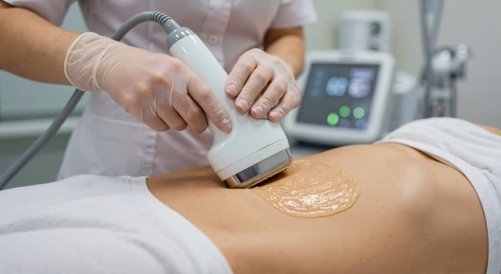 A medical professional using a handheld device for a treatment on a client's abdomen covered with a gel. The practitioner is wearing gloves, and a monitor is visible in the background, indicating a clinical environment.