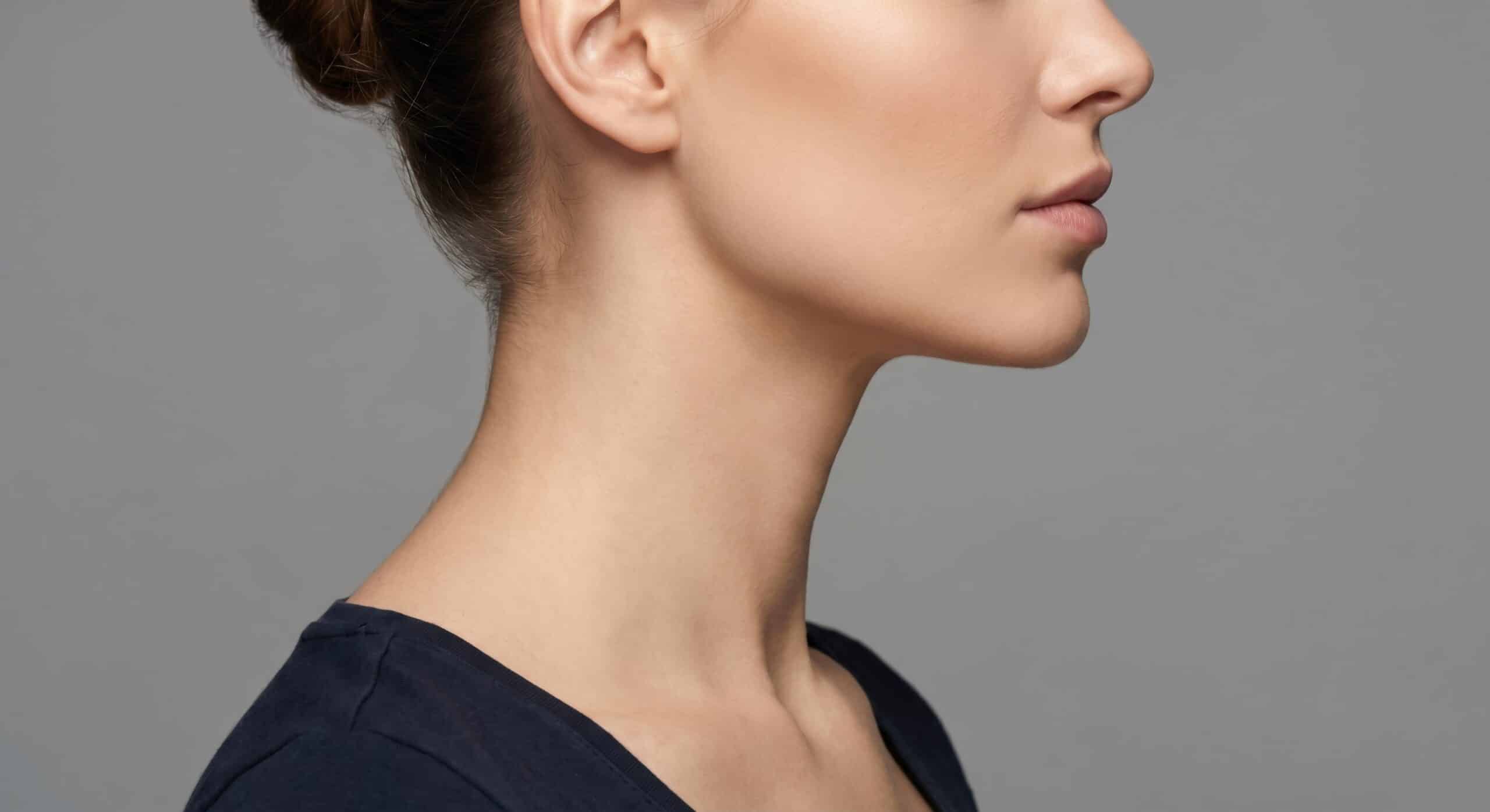 A profile view of a woman with a smooth complexion and a neat bun hairstyle, wearing a dark shirt. The background is a soft gray, emphasizing her facial features and neckline.