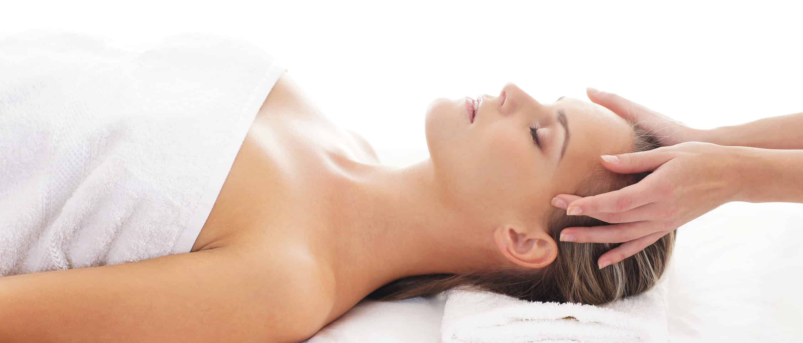 A woman lying on a massage table with her eyes closed, receiving a head massage. She is covered with a white towel, and her hair is pulled back. Soft white towels are visible in the background, creating a calming atmosphere.