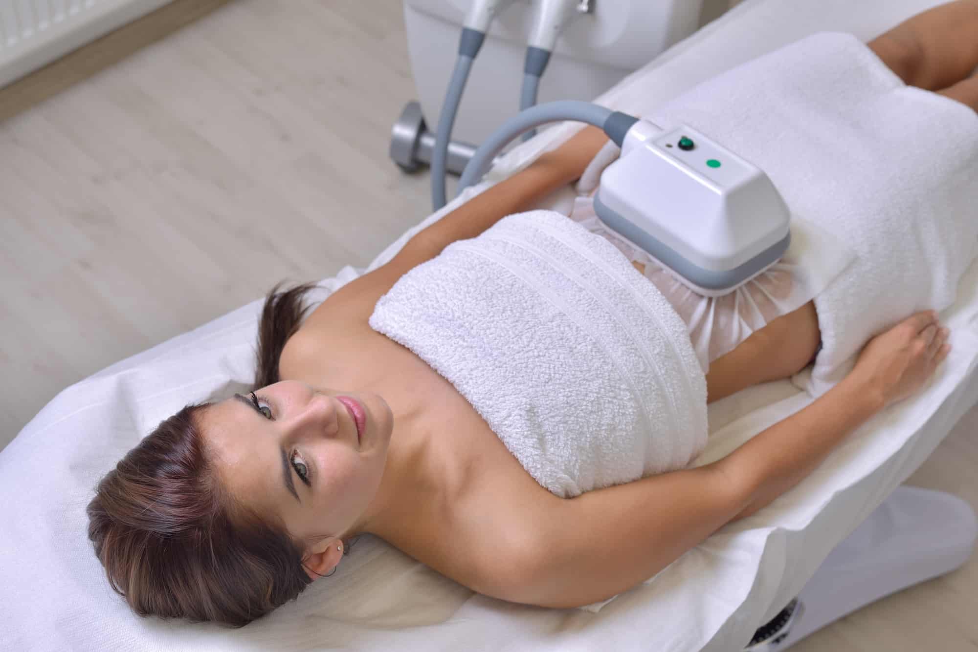 A woman lying on a treatment table covered with a white towel. She has a device positioned on her abdomen, and she is looking up towards the camera. The room has a modern appearance with clinical equipment visible in the background.